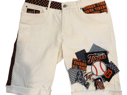 A pair of off-white denim shorts featuring a Texas Rangers baseball-themed patchwork graphic on the right leg and a patterned orange-and-black waistband, displayed against a plain white background.