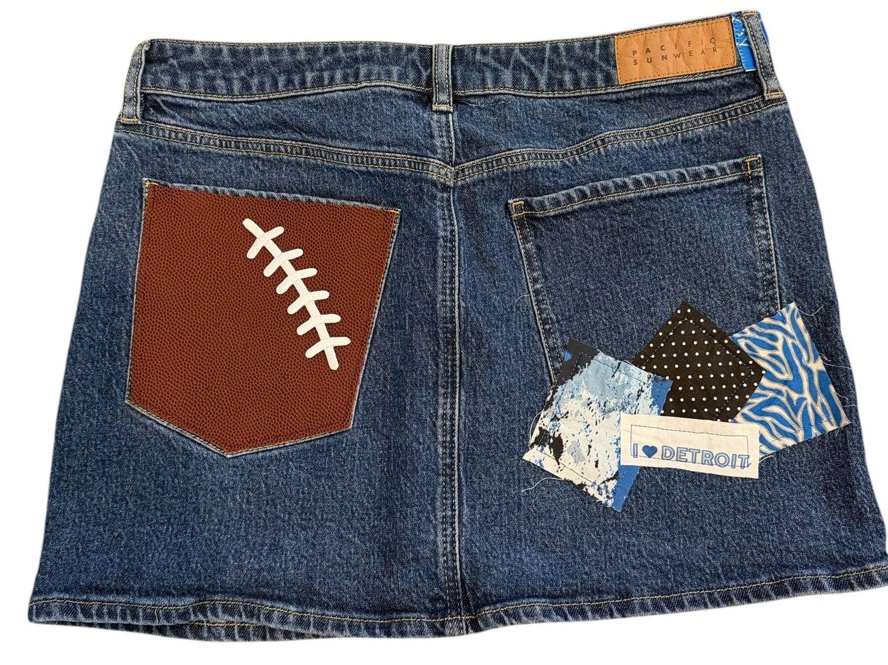 A pair of blue denim jeans laid flat on a white background, featuring a brown football-shaped patch on the back pocket with white lacing details, a patterned fabric patch on the front pocket, and a small tan brand label.