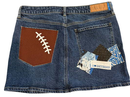 A pair of blue denim jeans laid flat on a white background, featuring a brown football-shaped patch on the back pocket with white lacing details, a patterned fabric patch on the front pocket, and a small tan brand label.
