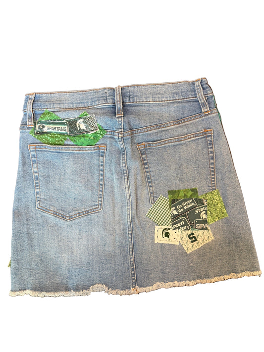 Light blue denim mini skirt with frayed hems, featuring a green and white embroidered patch on the left back pocket and a multicolor patch on the right back pocket.